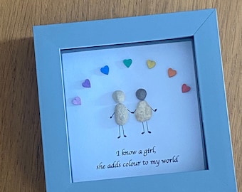 May include: A framed artwork with a white background featuring two figures made of stones, holding hands. The figures are surrounded by colorful hearts. The text "I know a girl, she adds colour to my world" is written below the figures.