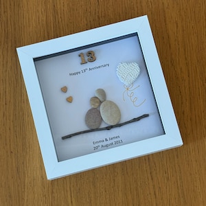 May include: A white framed picture with a white background. The picture features a wooden number 13, the words "Happy 13th Anniversary", a small twig with a heart-shaped pebble decoration, and the text "Emma & James 20th August 2011".