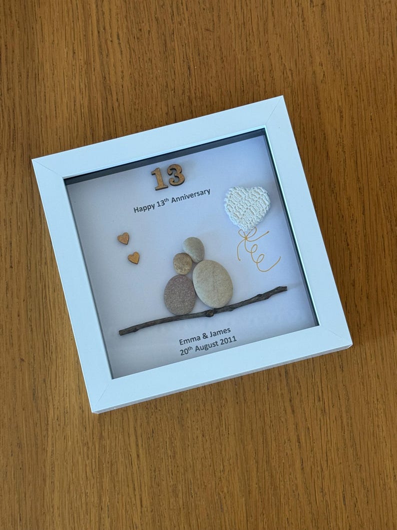 May include: A white framed picture with a white background. The picture features a wooden number 13, the words "Happy 13th Anniversary", a small twig with a heart-shaped pebble decoration, and the text "Emma & James 20th August 2011".