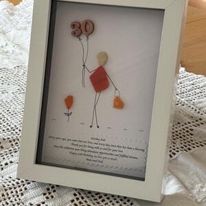 May include: A white framed picture with a hand-drawn illustration of a person holding balloons that say "30". The person is made of colorful sea glass. The picture includes a handwritten message that says "Thirty years ago, you came into our lives, and every day since then has been a blessing. Thank you for being with us and for your love. May this milestone year bring abundant opportunities and fulfilled dreams. Happy 30th birthday, we love you so much. Mom and Dad."