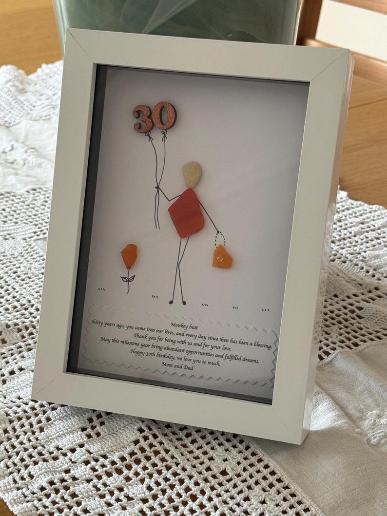 May include: A white framed picture with a hand-drawn illustration of a person holding balloons that say "30". The person is made of colorful sea glass. The picture includes a handwritten message that says "Thirty years ago, you came into our lives, and every day since then has been a blessing. Thank you for being with us and for your love. May this milestone year bring abundant opportunities and fulfilled dreams. Happy 30th birthday, we love you so much. Mom and Dad."