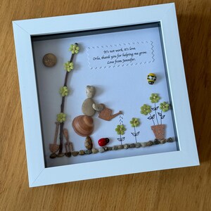 Puede incluir: Una obra de arte enmarcada que presenta a una persona de guijarros regando flores con una regadera. La obra de arte est&aacute; hecha con materiales naturales e incluye un mensaje escrito a mano que dice: "No es trabajo, es amor. Orla, gracias por ayudarme a crecer. Con cari&ntilde;o, Jennifer."