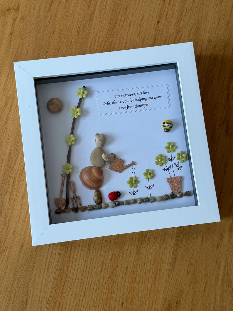Puede incluir: Una obra de arte enmarcada que presenta a una persona de guijarros regando flores con una regadera. La obra de arte est&aacute; hecha con materiales naturales e incluye un mensaje escrito a mano que dice: "No es trabajo, es amor. Orla, gracias por ayudarme a crecer. Con cari&ntilde;o, Jennifer."