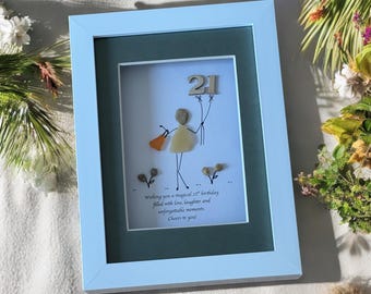May include: A framed pebble art piece with a stick figure holding a balloon that says "21". The text below reads "Wishing you a magical 21st birthday filled with love, laughter and unforgettable moments. Cheers to you!"