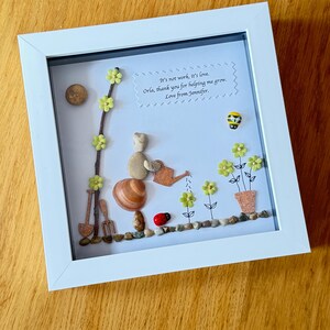Puede incluir: Una obra de arte enmarcada hecha con piedras y otros materiales naturales. La obra representa a una persona regando flores con una regadera. El texto de la imagen dice "It's not work, it's love. Orla, thank you for helping me grow. Love from Jennifer."