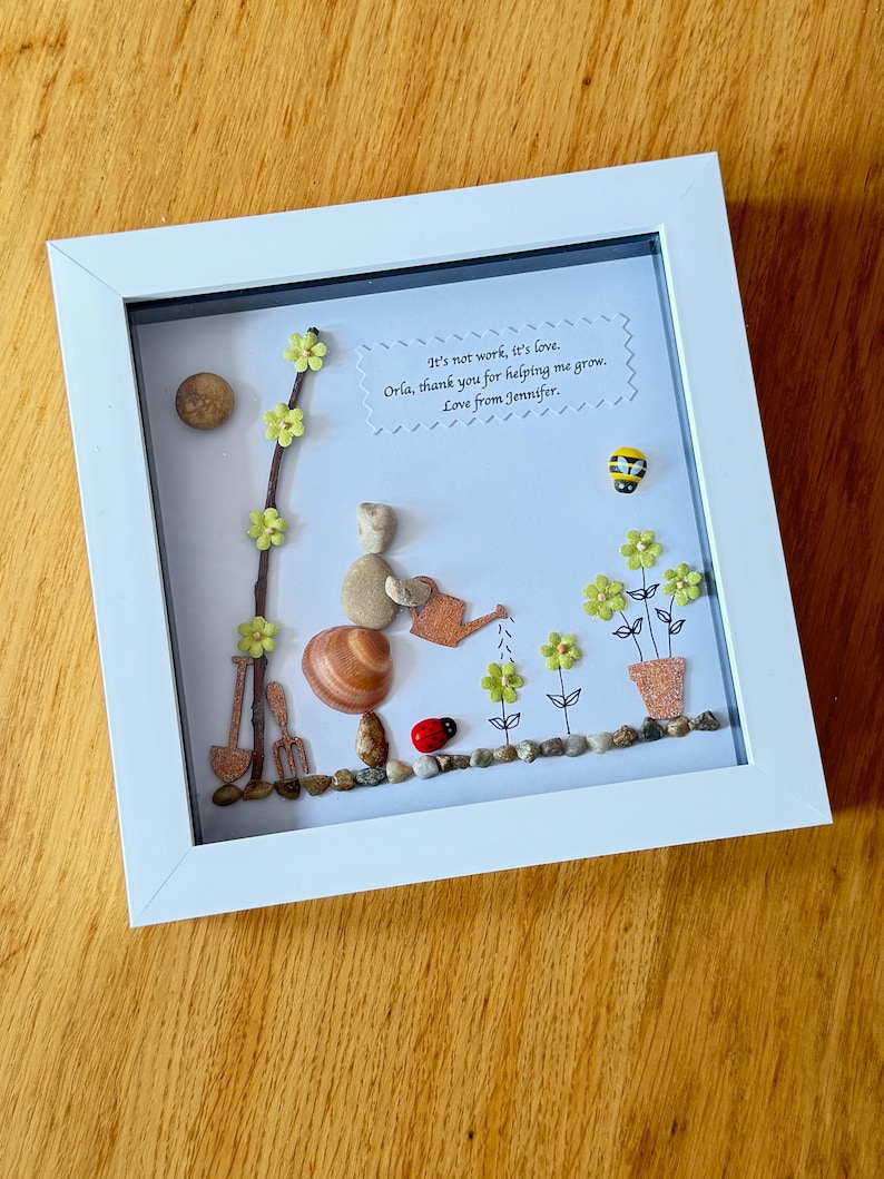 Puede incluir: Una obra de arte enmarcada hecha con piedras y otros materiales naturales. La obra representa a una persona regando flores con una regadera. El texto de la imagen dice "It's not work, it's love. Orla, thank you for helping me grow. Love from Jennifer."