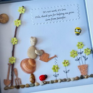 Puede incluir: Una obra de arte enmarcada hecha con materiales naturales, que representa a una persona hecha de piedras regando flores. Las flores est&aacute;n hechas de papel y purpurina, y la persona sostiene una regadera de madera. La obra de arte incluye el texto "It's not work, it's love. Orla, thank you for helping me grow. Love from Jennifer."