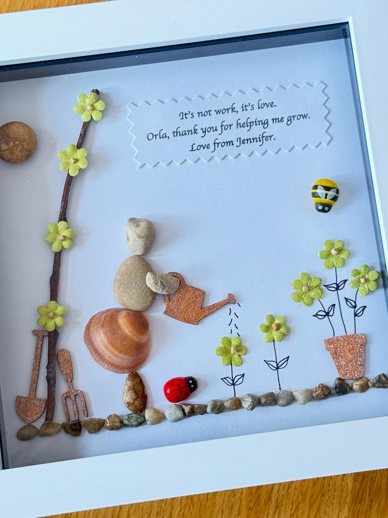 Puede incluir: Una obra de arte enmarcada hecha con materiales naturales, que representa a una persona hecha de piedras regando flores. Las flores est&aacute;n hechas de papel y purpurina, y la persona sostiene una regadera de madera. La obra de arte incluye el texto "It's not work, it's love. Orla, thank you for helping me grow. Love from Jennifer."