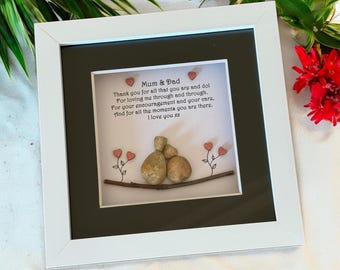 Puede incluir: Una pieza de arte de guijarros enmarcada con un mensaje para mamá y papá. El mensaje dice "Gracias por todo lo que sois y hacéis. Por quererme a través del tiempo. Por vuestro ánimo y vuestros cuidados. Y por todos los momentos en que estáis ahí. Os quiero xx". El arte de guijarros representa dos figuras con corazones a su alrededor.