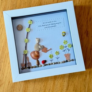 Puede incluir: Una pieza de arte de guijarros enmarcada con un mensaje que dice: "No es trabajo, es amor. Orla, gracias por ayudarme a crecer. Con amor, Jennifer." El arte representa a una persona regando flores con una regadera, una mariquita y una abeja.