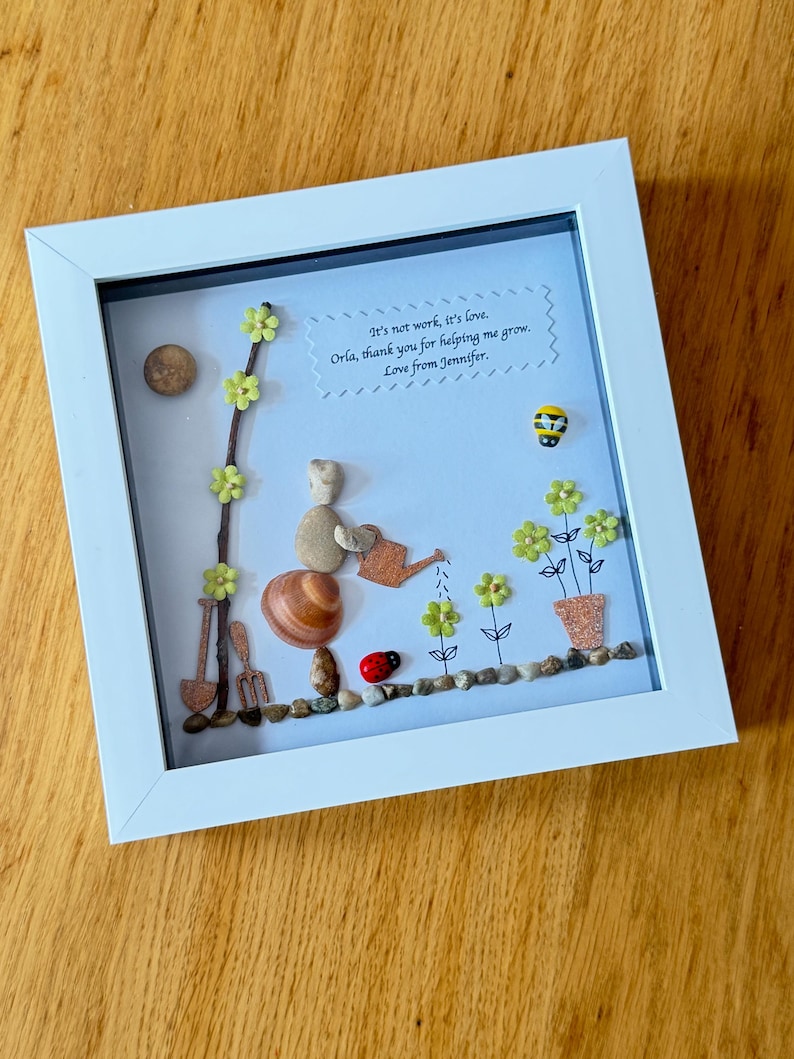 Puede incluir: Una pieza de arte de guijarros enmarcada con un mensaje que dice: "No es trabajo, es amor. Orla, gracias por ayudarme a crecer. Con amor, Jennifer." El arte representa a una persona regando flores con una regadera, una mariquita y una abeja.