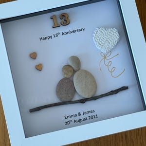 May include: A white frame with a white background. The number 13 is made of wood and is in the top left corner. The text "Happy 13th Anniversary" is below the number. A white heart with a gold string is in the top right corner. A twig with a small pile of stones is in the bottom center. The text "Emma & James 20th August 2011" is below the twig.