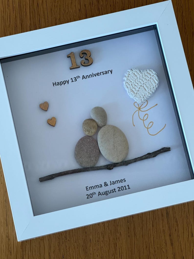 May include: A white frame with a white background. The number 13 is made of wood and is in the top left corner. The text "Happy 13th Anniversary" is below the number. A white heart with a gold string is in the top right corner. A twig with a small pile of stones is in the bottom center. The text "Emma & James 20th August 2011" is below the twig.