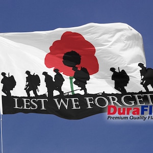 May include: A white flag featuring a red poppy and black silhouettes of soldiers. The words "LEST WE FORGET" are written in white on a black banner. The flag is displayed against a blue sky, a symbol of remembrance.