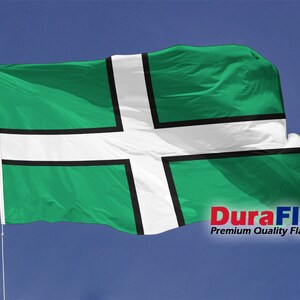 Può includere: Una bandiera verde e bianca con un disegno a croce, sventolante nel vento contro un cielo blu. La bandiera presenta una croce bianca con bordi neri su sfondo verde. Le parole "DuraFlag" e "Premium Quality Flags" sono visibili.