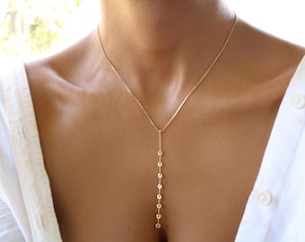 May include: A delicate gold necklace with a vertical bar pendant. The pendant features a series of small, round, clear gemstones. The necklace is worn against a person's skin, with a white shirt visible in the background.