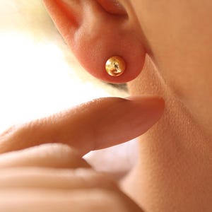 May include: A small, round, gold-colored stud earring is visible on an ear. The earring is a simple, polished sphere. A finger gently touches the ear, near the earring. The skin tone is light.