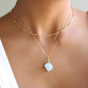 Könnte beinhalten: Eine goldene Kettenkette mit einem hellblauen Edelstein-Anhänger. Die Kette ist mit einer zweiten, dünneren goldenen Kette geschichtet.