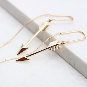 May include: A pair of gold arrow earrings with delicate chains. The arrows are detailed with lines and have a pointed tip.