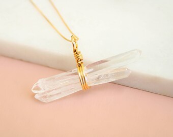 May include: A gold-colored necklace featuring two clear quartz crystal points. The crystals are wrapped together with gold wire and suspended from a delicate gold chain. The background is a soft pink and white.