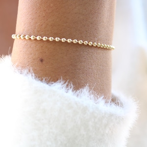 May include: A delicate gold ball chain bracelet is worn on the wrist. The bracelet is composed of small, evenly spaced spheres. The wrist is against a white, fuzzy sweater.