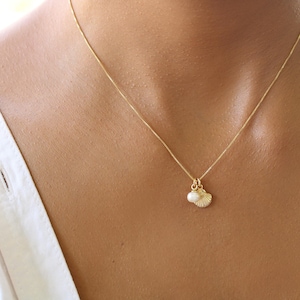 May include: A delicate gold necklace featuring a small pendant with a seashell design and a pearl. The necklace is worn against a person's skin, with a white shirt visible in the background. The pendant is a small, elegant piece of jewelry.