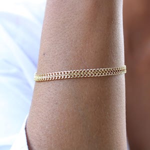May include: A gold-colored bracelet with a double chain design. The bracelet is worn on the arm and appears to be made of metal. The jewelry is a simple yet elegant accessory.