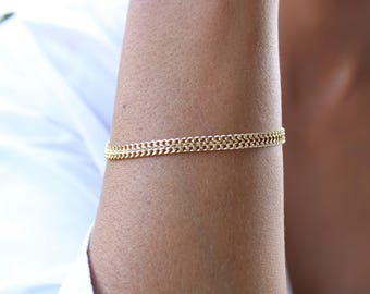 Puede incluir: Una pulsera dorada con un diseño de doble cadena. La pulsera se lleva en el brazo y parece ser de metal. La joya es un accesorio sencillo pero elegante.