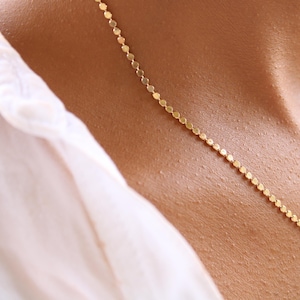 Puede incluir: Un delicado collar de oro con un diseño de discos repetidos. El collar se lleva sobre la piel, con una camisa blanca visible en el fondo. El collar es una joya sencilla y elegante.