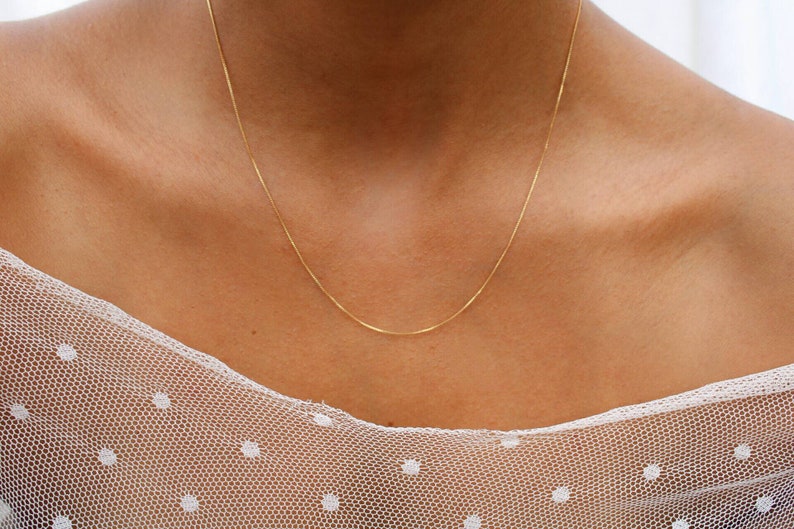 May include: A simple gold chain necklace on a person wearing a white dotted mesh top.