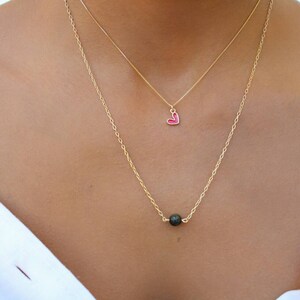 Pu&ograve; includere: Una collana con catena dorata con un piccolo ciondolo a forma di cuore rosso e una perla di lava nera.