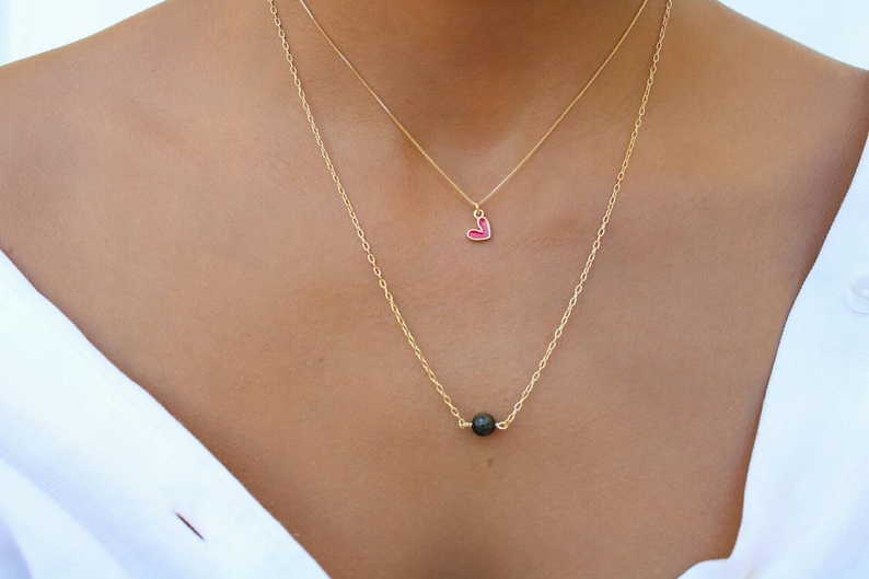 Pu&ograve; includere: Una collana con catena dorata con un piccolo ciondolo a forma di cuore rosso e una perla di lava nera.