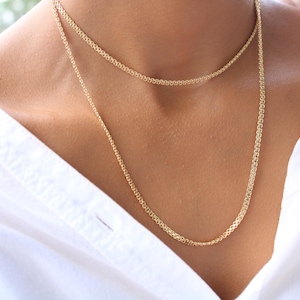 May include: Two delicate gold necklaces of different lengths are displayed against a white shirt. The necklaces have a fine chain design, with a subtle texture. The jewelry is a warm gold colour, creating a simple yet elegant look.