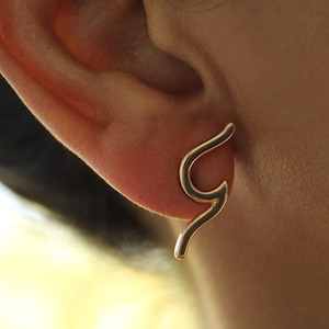 May include: A close-up of a gold-colored earring in the shape of a curved line. The earring is worn on an ear, with the ear and surrounding skin visible. The earring has a smooth, polished surface and a minimalist design.