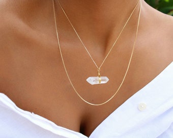 May include: A gold chain necklace with a clear quartz crystal pendant. The necklace is layered with a second, thinner gold chain.