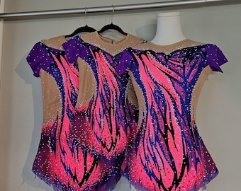 May include: Three rhythmic gymnastics leotards. Each leotard features a nude-colored bodice with a vibrant design of pink, purple, and blue accents. The design includes a pattern of flowing lines and is embellished with sparkling rhinestones. The leotards hang on a clothing rack.
