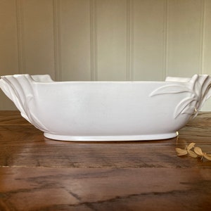 May include: White ceramic serving bowl with a decorative leaf design on the sides. The bowl is oval shaped and has a smooth, glossy finish.