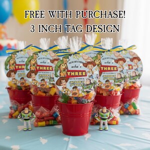 May include: Several red buckets filled with candy and Toy Story-themed party favors. Each favor bag has a circular tag with the text "THREE" and character illustrations. The image includes the text "FREE WITH PURCHASE! 3 INCH TAG DESIGN".