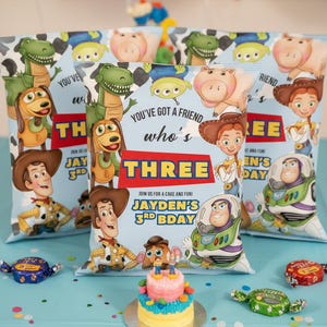 May include: Toy Story themed party favors with character illustrations. The favors are light blue with the text "You've got a friend who's THREE". A small cake and wrapped candies are on a blue tablecloth. Balloons are in the background.