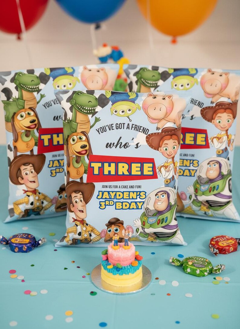 May include: Toy Story themed party favors with character illustrations. The favors are light blue with the text "You've got a friend who's THREE". A small cake and wrapped candies are on a blue tablecloth. Balloons are in the background.