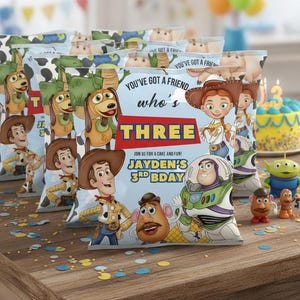 May include: Several light blue party favor bags featuring Toy Story characters, with the text "YOU'VE GOT A FRIEND who's THREE JAYDEN'S 3RD BDAY". The bags are on a wooden table with confetti and a birthday cake.
