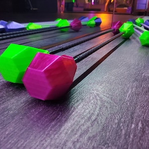 May include: A collection of colorful, geometric-shaped objects with long, dark handles. The objects are in shades of green, pink, and blue. The objects are arranged on a dark, textured surface, with reflections visible.
