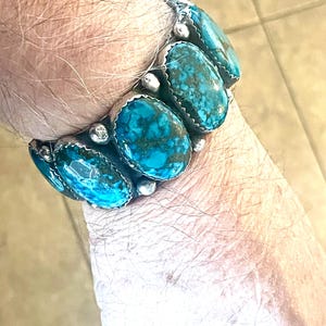 Può includere: Un braccialetto di turchese con dettagli argentati. Il braccialetto presenta diverse pietre di turchese ovali, ciascuna incastonata in una lunetta d'argento. Le pietre mostrano una vivace tonalità blu-verde con motivi a matrice scura. Il braccialetto è indossato al polso.