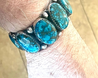 May include: A turquoise bracelet with silver detailing. The bracelet showcases several oval turquoise stones, each set in a silver bezel. The stones display a vivid blue-green hue with dark matrix patterns. The bracelet is worn on a wrist.