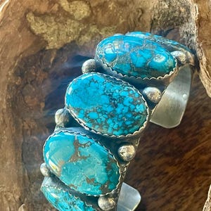 Könnte beinhalten: Ein silbernes Armreif-Armband mit vier großen, ovalen Türkissteinen. Die Steine haben eine leuchtende türkise Farbe mit braunen Matrixmustern. Das Armband hat ein rustikales, handgefertigtes Aussehen mit silbernen Akzenten.