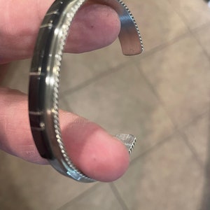 May include: A silver cuff bracelet with a beaded edge and a dark, possibly inlaid, central band. The bracelet is open and held by a hand, showcasing its design and craftsmanship. The metal appears to be polished.
