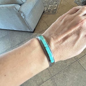 Puede incluir: Brazalete plateado con incrustaciones rectangulares de turquesa. El brazalete tiene un diseño de borde trenzado. Las piedras turquesas son de color azul verdoso brillante. El brazalete se lleva en la muñeca.