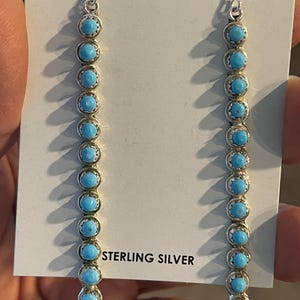 May include: Sterling silver dangle earrings featuring a series of round, light blue turquoise stones. The stones are set in a silver bezel setting and hang from a silver hook. The earrings are displayed on a white card with the text "STERLING SILVER".