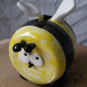 May include: A ceramic bee-shaped money box with black and yellow stripes and white accents. The bee has large, white eyes and a black nose. The money box is sitting on a wooden surface with several British coins in front of it.