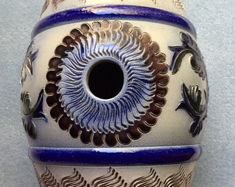 May include: A white ceramic barrel-shaped container with blue and gold painted designs. The container has a round hole in the center and is decorated with floral patterns.